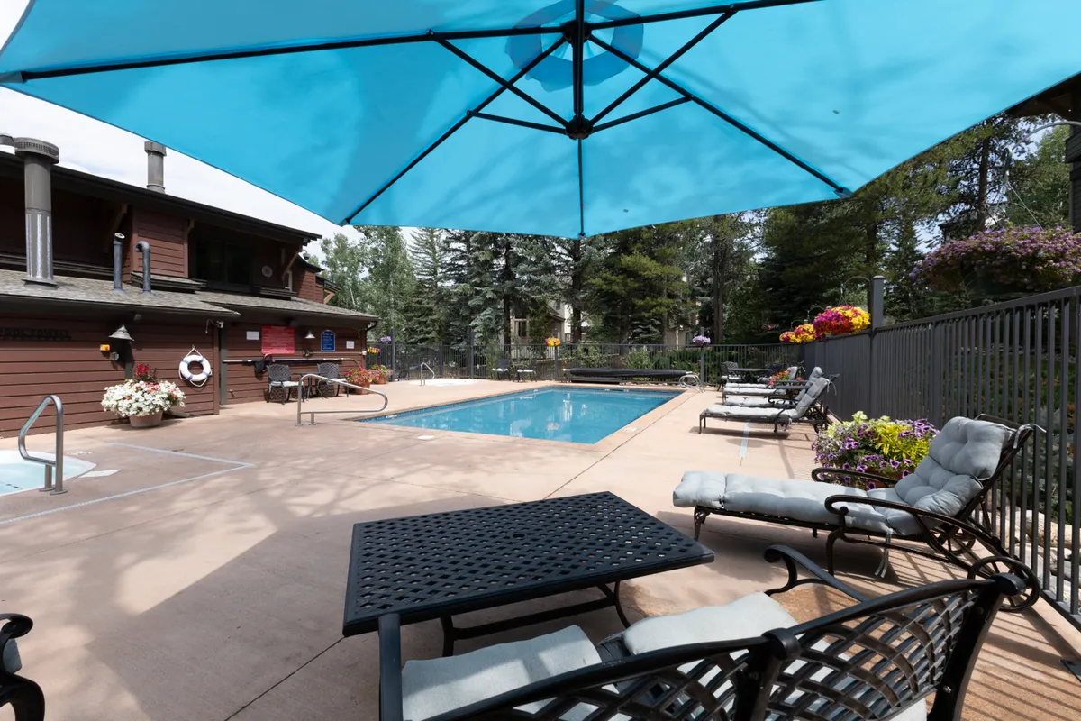 A wider exterior shot featuring a sparkling swimming pool bordered by an outdoor seating area with a large umbrella and lush plantings. The spacious layout encourages group activities, poolside barbecues, and socializing in a relaxed, welcoming atmosphere.