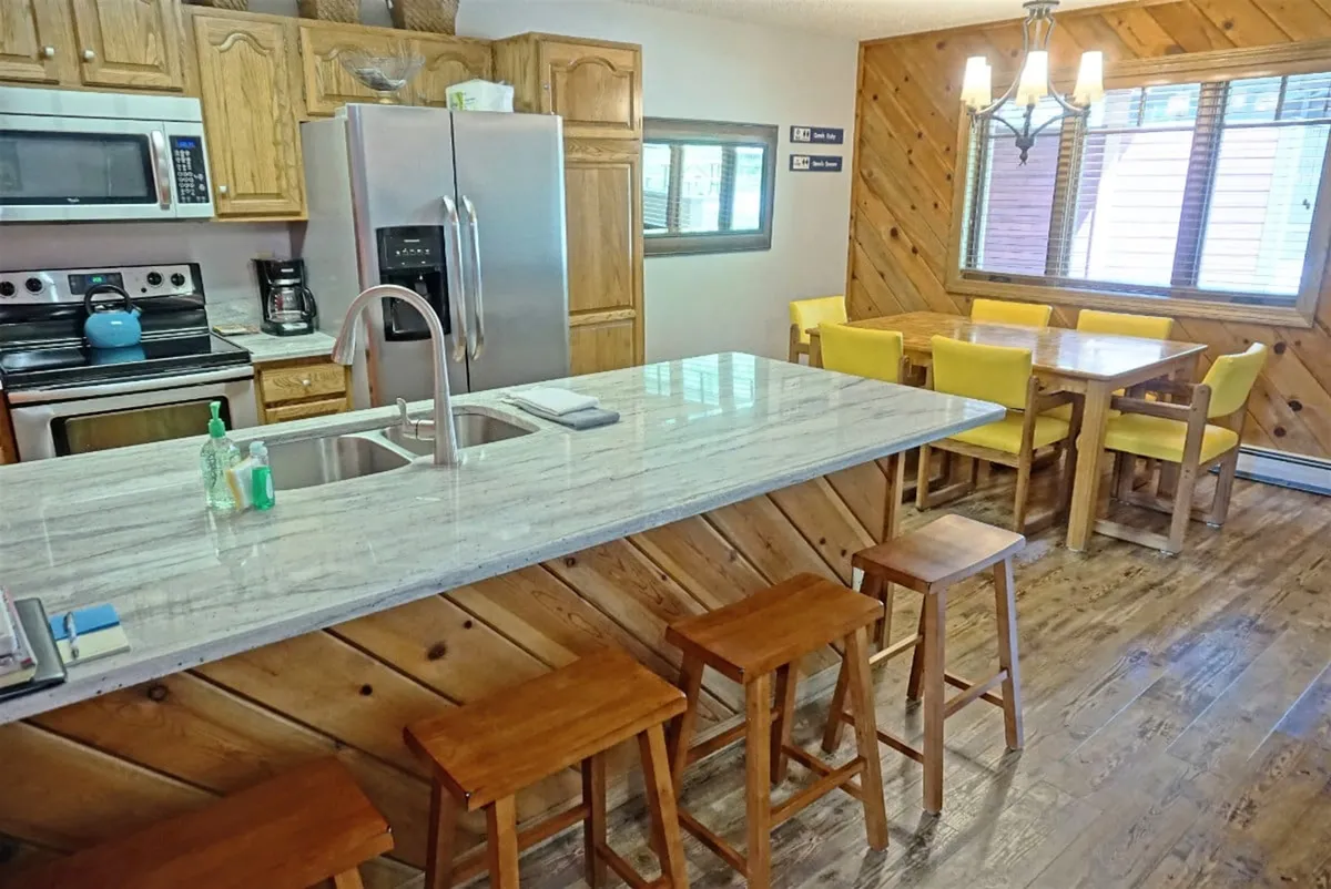A warmly lit kitchen and dining area with a central island, stainless steel appliances, a family-sized dining table, and wooden stools. The setting suggests a hub for home-cooked meals, casual gatherings, and shared celebrations.