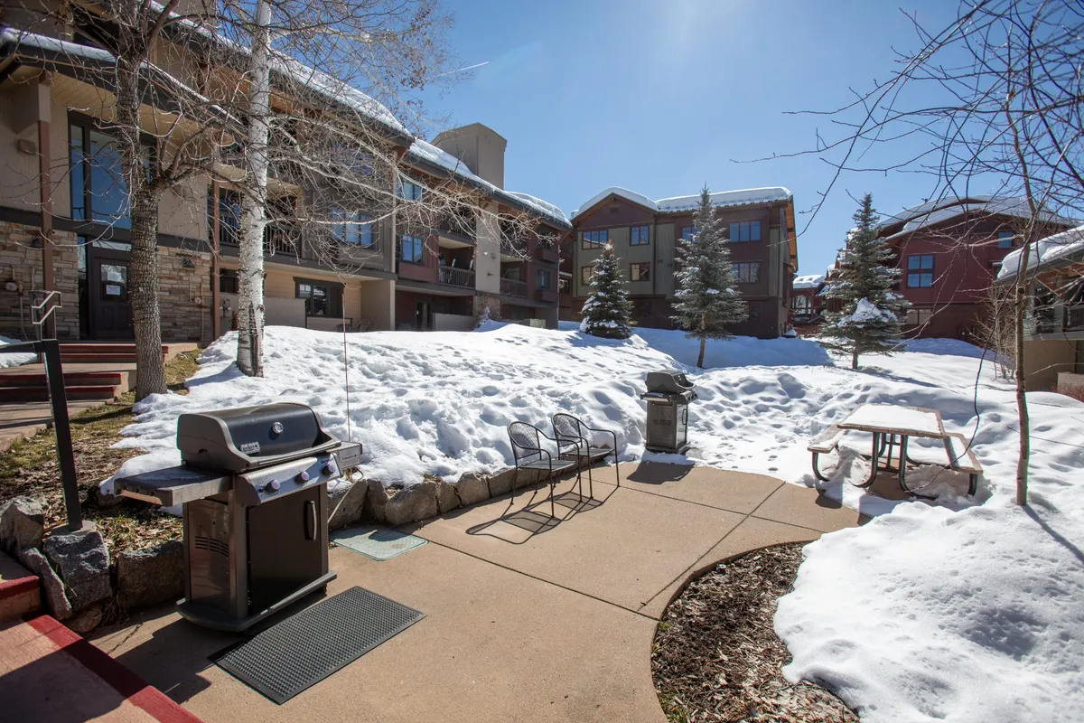 A snow-dusted outdoor area with a grill, picnic table, and view of the building's exterior and surrounding trees. The scene conveys wintertime fun and the convenience of direct yard access from the home.