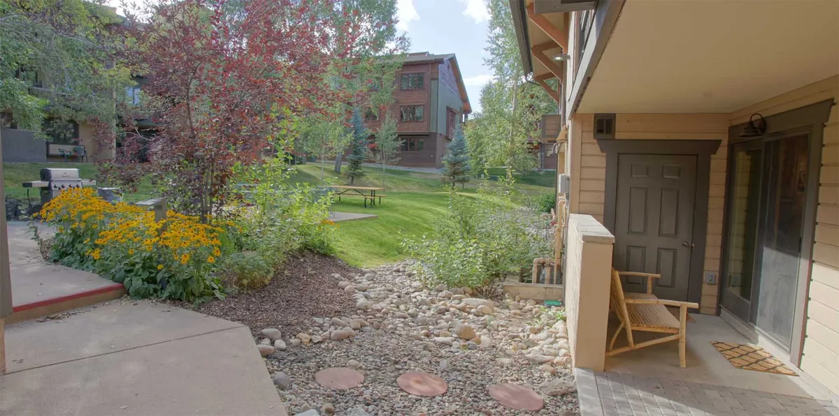 The exterior of the condo is framed by vibrant yellow flowers and a lush, well-manicured lawn. A comfortable seating area sits invitingly on the porch, connected by a stone pathway, suggesting a peaceful place to relax. The overall mood is tranquil and inviting, evoking the feeling of a mountain retreat surrounded by nature.