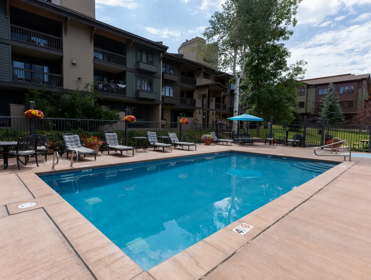 A vibrant exterior scene featuring a spacious swimming pool surrounded by lounge chairs, umbrellas, and decorative flower pots. The pool area is inviting, perfect for both socializing and relaxing, and is adjacent to the property’s building.