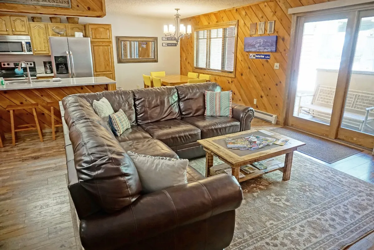 A wide view of the open-concept living area showing a plush leather sectional, a spacious dining area, a coffee table, and sliding glass doors that bring in natural light. The layout encourages easy movement and interaction among guests.