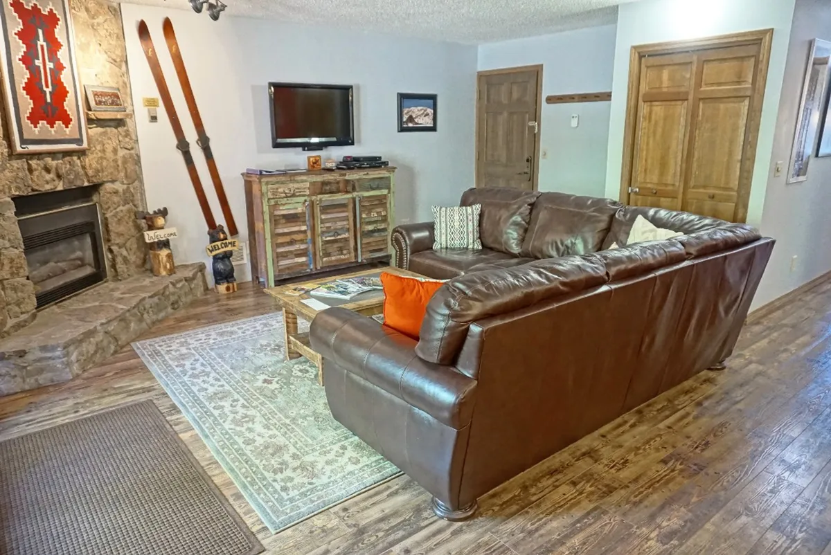 A spacious, rustic-themed interior with a stone fireplace, leather sofa, wooden furniture, decorative skis, and a TV. The setting suggests leisure time spent entertaining or relaxing after outdoor pursuits.