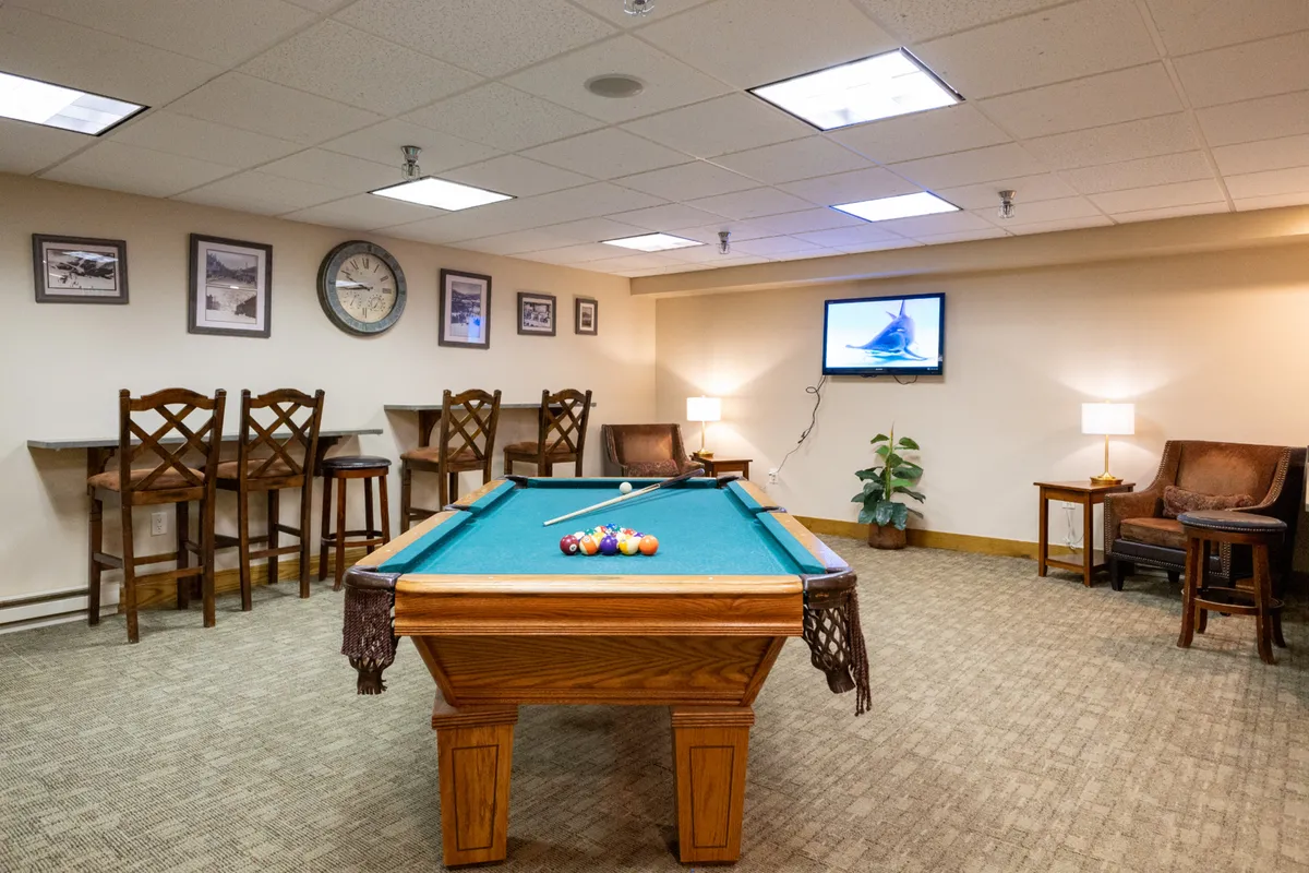 A stylish clubhouse interior featuring a pool table, bar stools, a television, and a comfortable seating area. The space is modern and social, perfect for entertaining, relaxing, or playing games with friends and family.