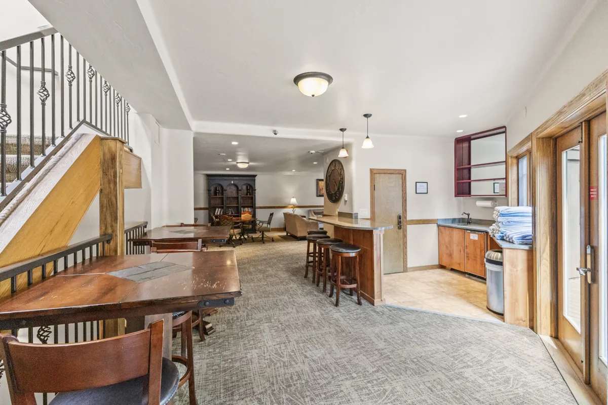 A welcoming entryway and gathering space featuring sturdy wooden tables, bar stools, a visible staircase leading up, and an entrance area with storage. The layout hints at easy transitions from outdoors to indoors, with ample space for gear drop-off and group organization.