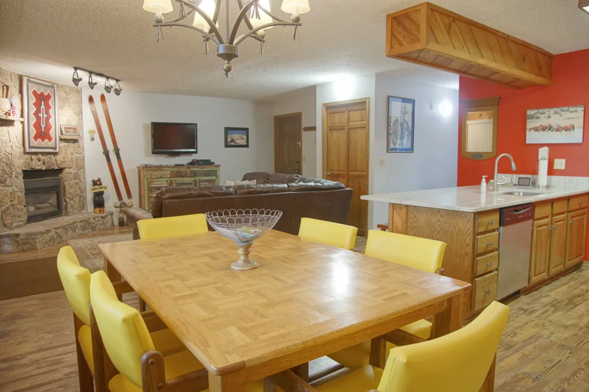 A warm, inviting dining area featuring a large table, a cozy fireplace, open kitchen, decorative skis, and a chandelier. The space is clearly designed for shared meals and memorable gatherings.
