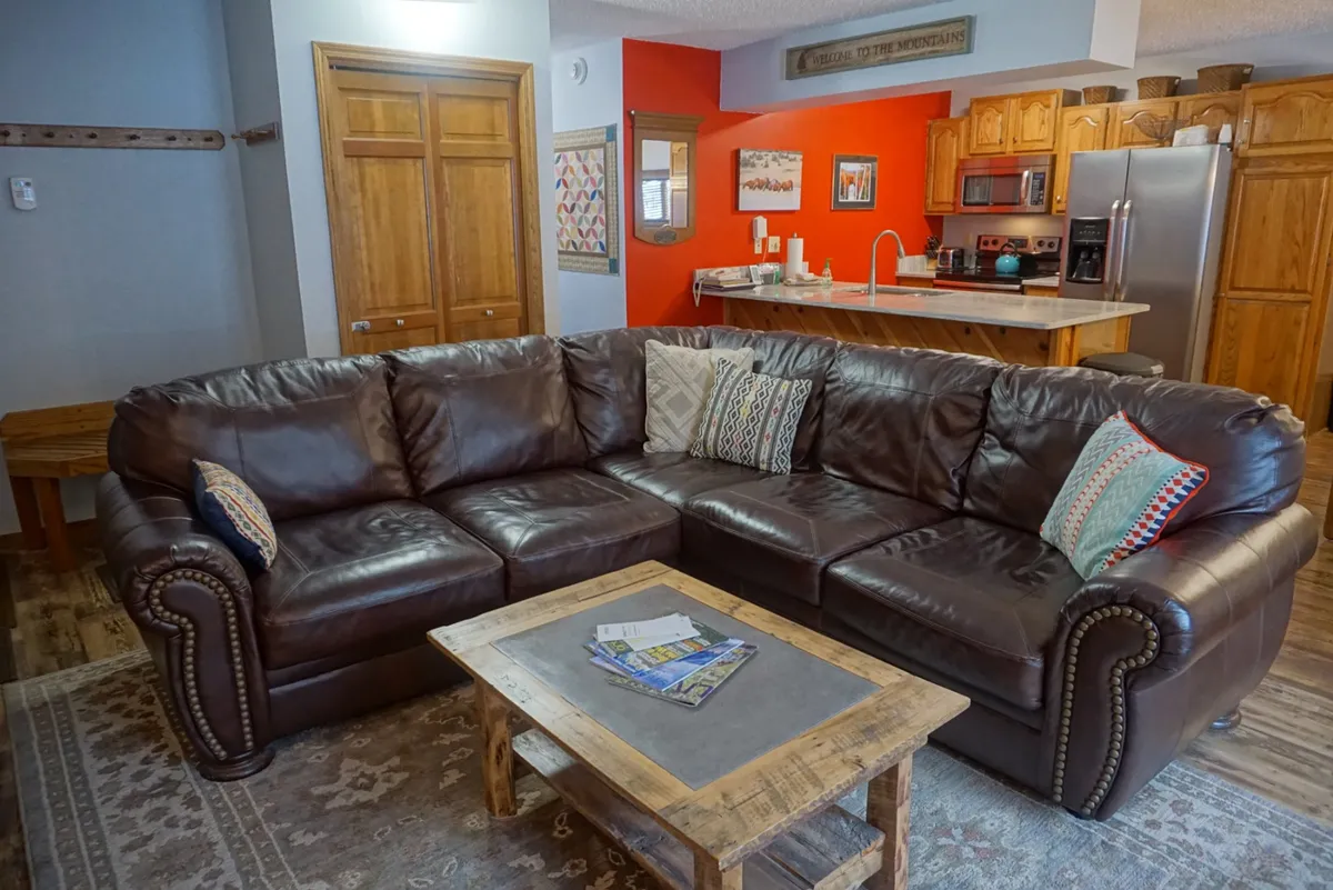 A cozy living space with a brown leather sectional adorned with colorful throw pillows, a wooden coffee table, and a view into the kitchen area in the background. The setup highlights both relaxation and the social connection between kitchen and living room.