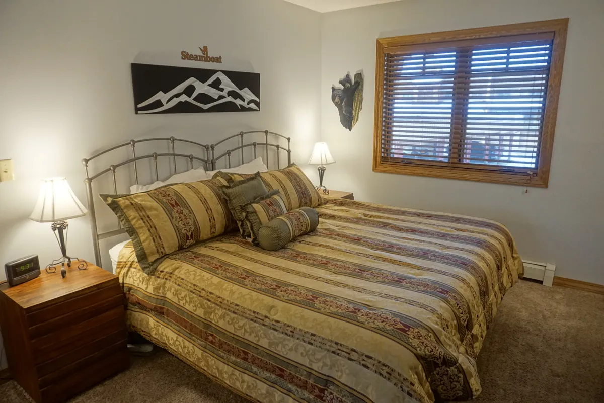 This image shows a bedroom with a plush bed adorned with decorative pillows, a nightstand with a lamp, and a wooden window with blinds letting in natural light. The overall mood is inviting and comfortable, with a cozy and restful atmosphere.