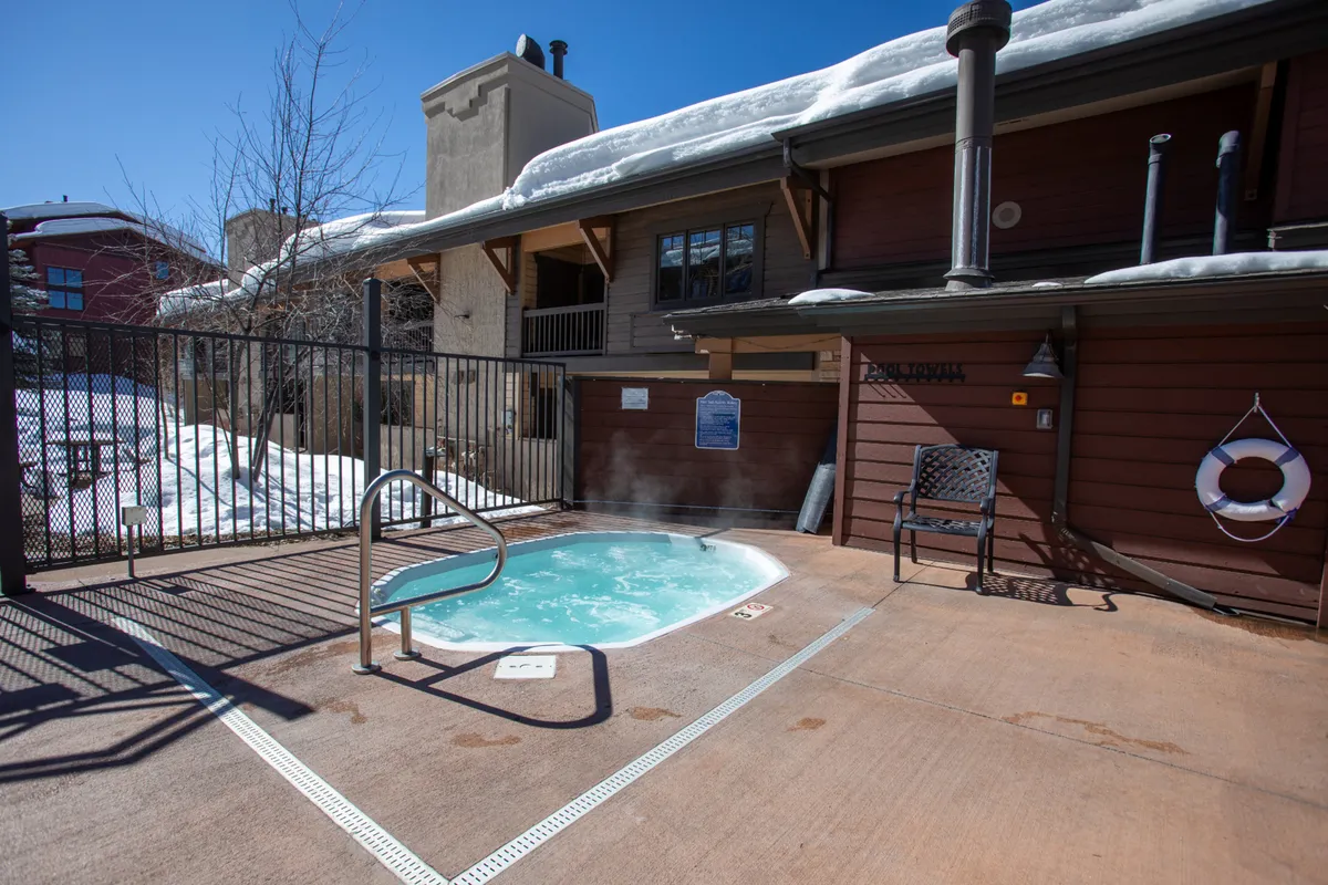 An inviting outdoor hot tub surrounded by a privacy fence and accompanied by a lounge chair, set in a tranquil, natural environment. The hot tub is large enough for a group and offers a relaxing spot to unwind, especially after a day of skiing or hiking.