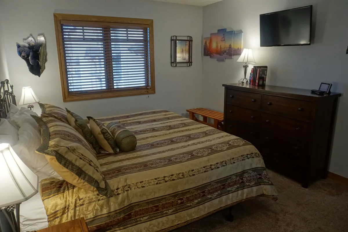A serene bedroom with a bed dressed in patterned bedding, decorative pillows, a dresser, window with blinds, soft table lamps, and wall art. The natural light and thoughtful decor create a tranquil retreat perfect for relaxing, reading, or enjoying sunset views.